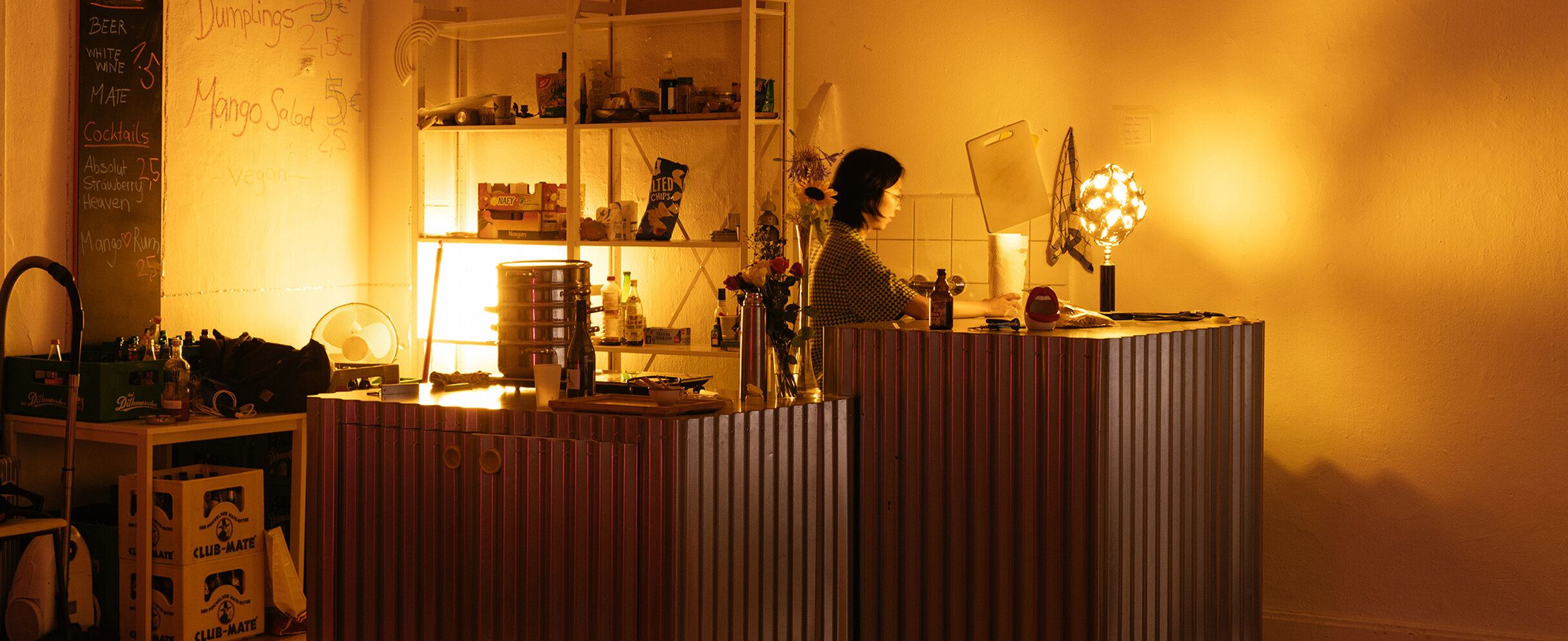 Foto: Eine Frau steht in einer Raumecke hinter einer Bar und wird von gelbem Licht angeleuchtet.